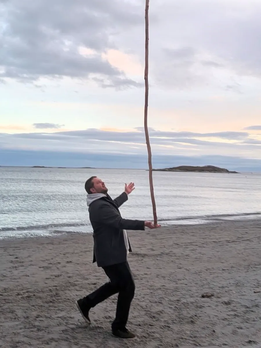 Brian balancing a long stick on a beach at dusk